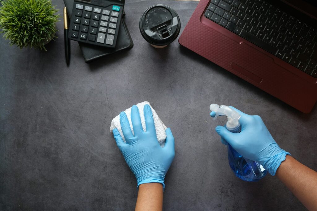 Blue-gloved hands cleaning a desk with a spray bottle